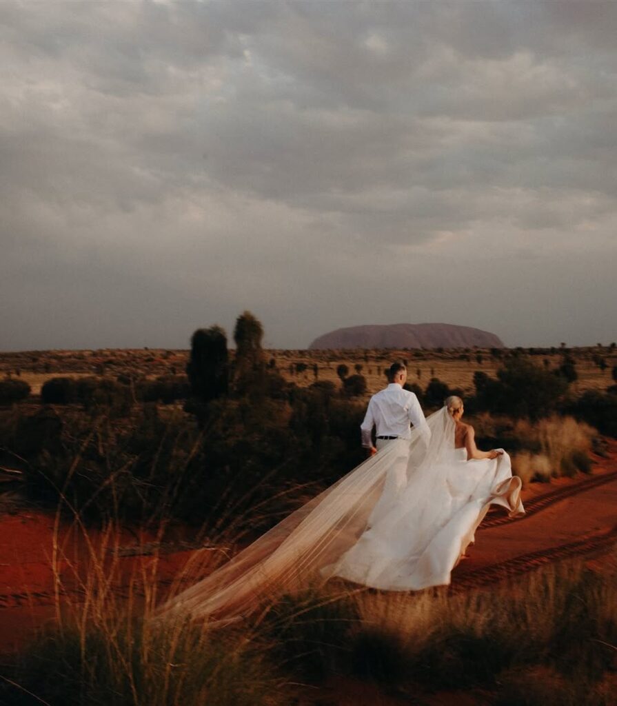 Where Sophistication Meets Storytelling: How Alysia Bridger brings love stories to life 4 Couple walking hand-in-hand in a scenic outback setting, bride in flowing wedding dress with long veil, showcasing Alysia Bridger's bespoke wedding design and storytelling.