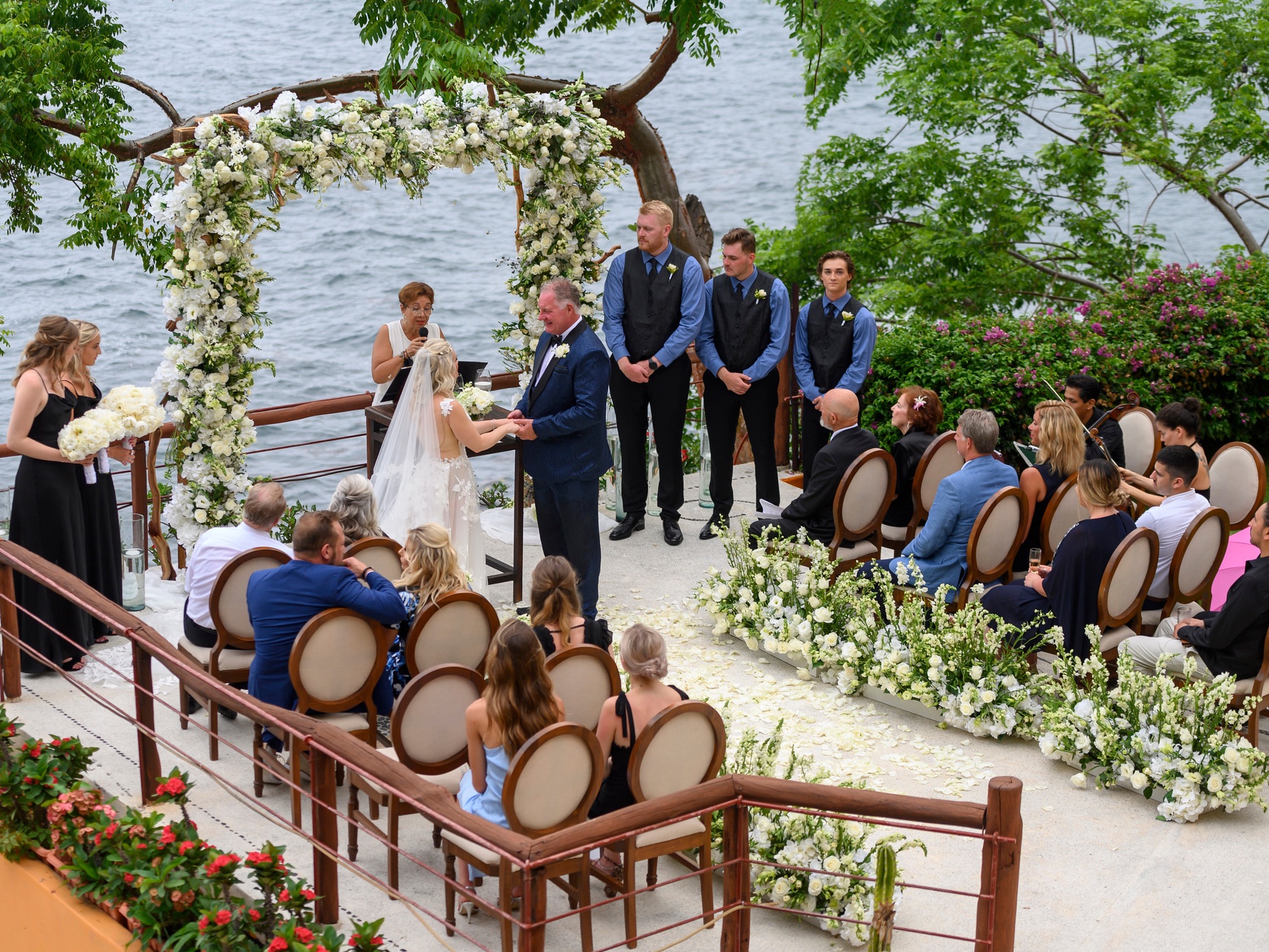 Wedding ceremony by the water, bride and groom exchanging vows under a floral arch, surrounded by guests in elegant attire, with lush greenery and floral arrangements enhancing the romantic setting.
