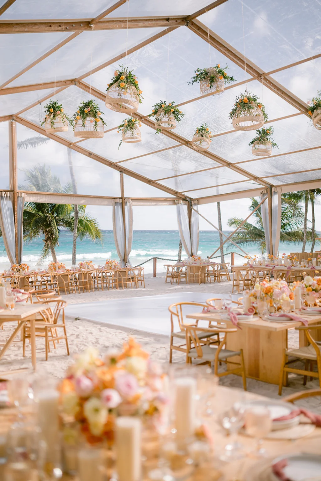 Elegant beachside wedding reception setup featuring floral centerpieces, wooden tables, and hanging floral arrangements under a clear tent, with ocean views in Tulum, Mexico.