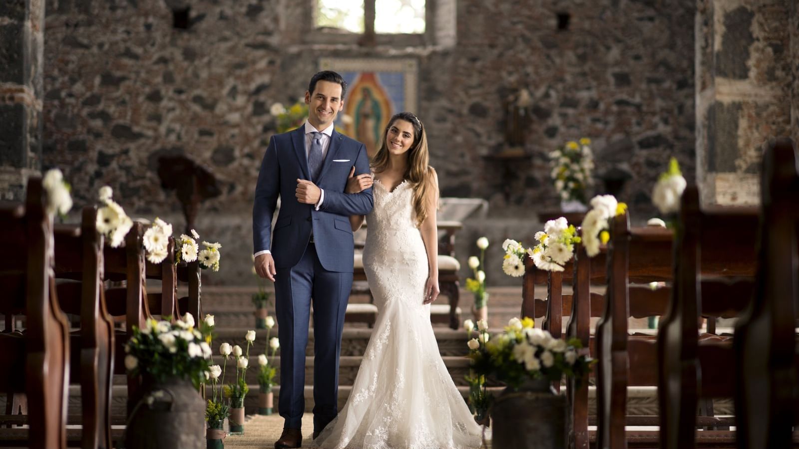 Bride and groom smiling together in a beautifully decorated wedding venue, featuring floral arrangements and rustic wooden pews, highlighting a romantic and elegant atmosphere for destination weddings in Mexico.