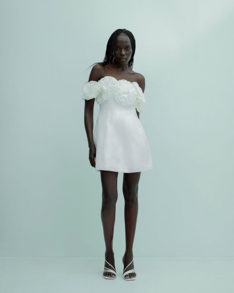 Model wearing a chic Acler mini dress with off-the-shoulder floral detailing, showcasing modern bridalwear aesthetics against a minimalist backdrop.