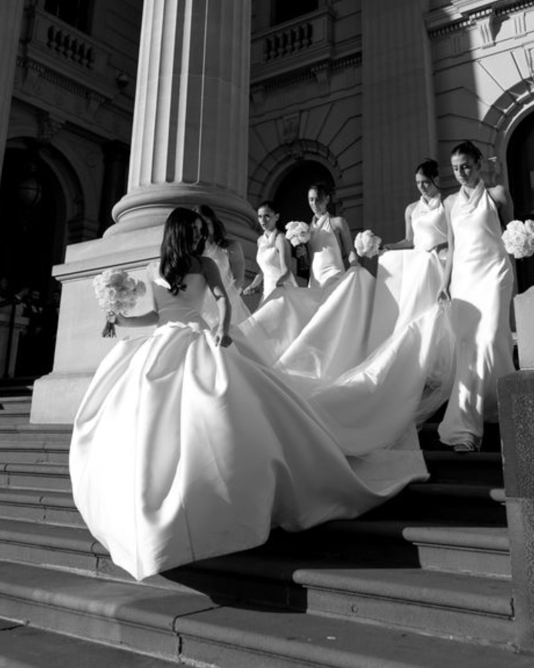 Bridesmaids in elegant white gowns descending grand stone steps, showcasing bridal fashion and wedding aesthetics.