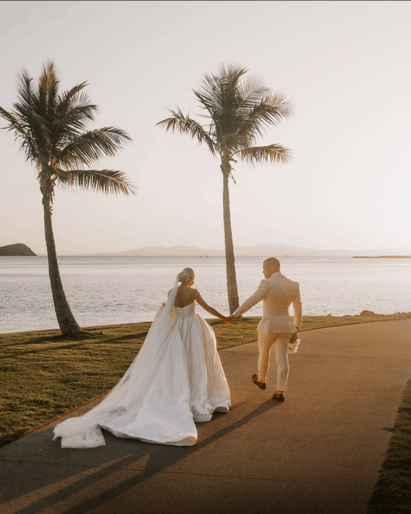 Couple walking hand in hand along a beachside path at sunset, surrounded by palm trees, showcasing the romantic setting of an Australian beach wedding.