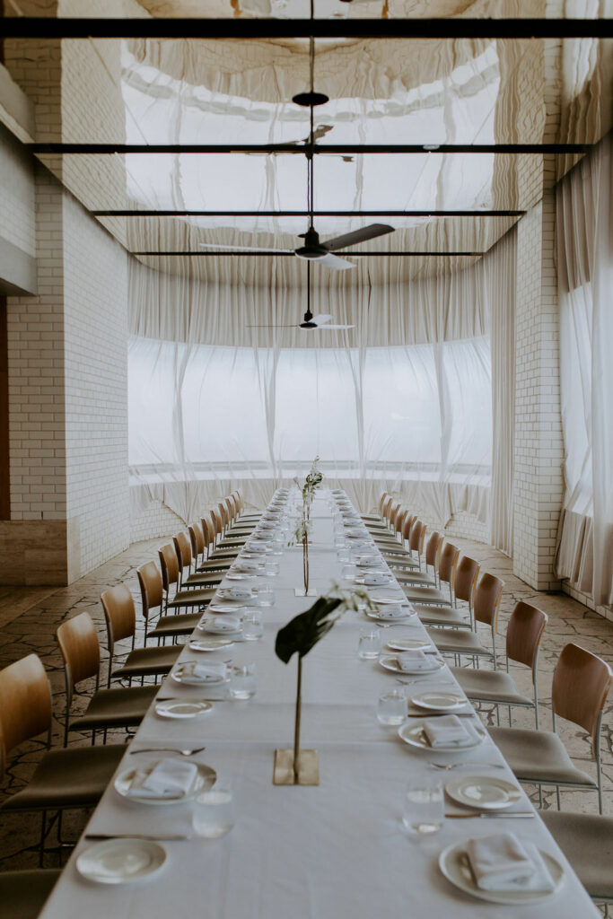 100 Breathtaking and Exclusive Australian Wedding Venues on every Couples Wish List! 58 Elegant dining setup in the Grand Room at The Calile Hotel, featuring a long table with white tablecloth, plates, glassware, and floral centerpieces, surrounded by large windows and soft drapery, ideal for sophisticated wedding receptions.
