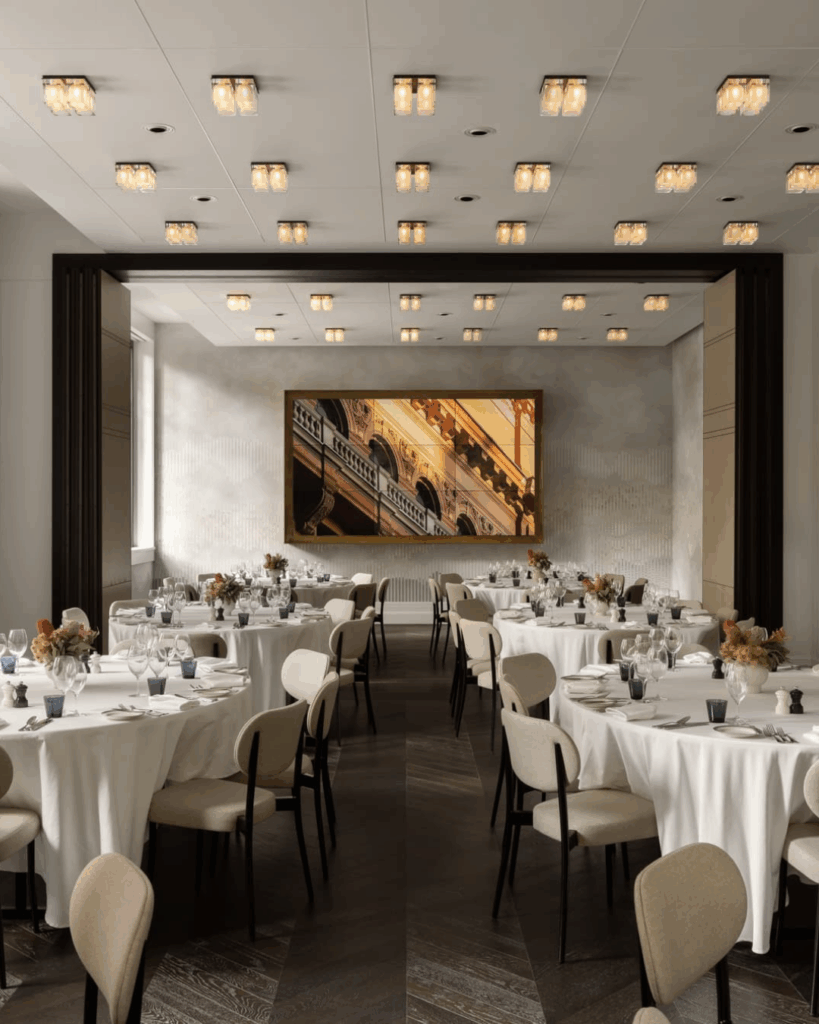 100 Breathtaking and Exclusive Australian Wedding Venues on every Couples Wish List! 2 Elegant dining room at Capella Sydney featuring round tables set with white tablecloths, glassware, and floral centerpieces, adorned with stylish lighting fixtures and a large artwork on the wall, ideal for luxurious wedding receptions.