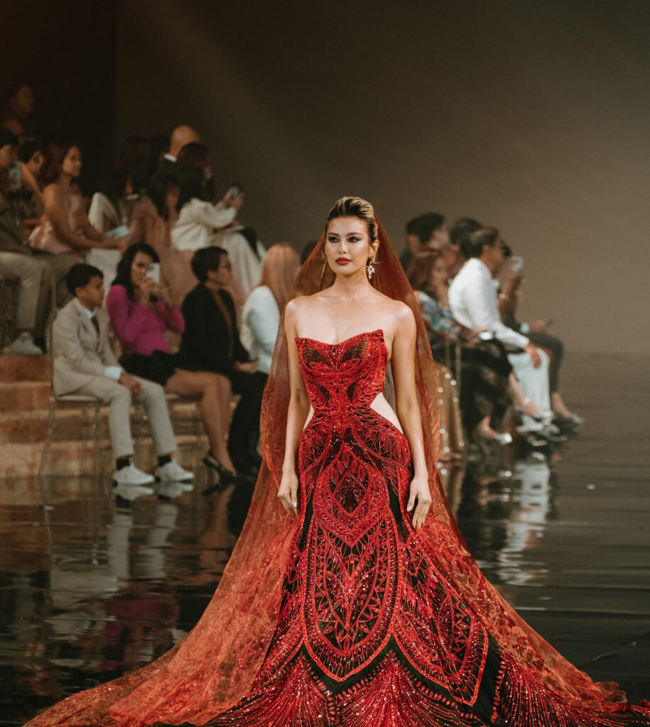 The World Has Been GIDEONIZED With A Bridal Fashion Extravaganza In The Philippines: Marry Me At Marriott X Gideon Hermosa 9 Model showcasing a stunning red and black bridal gown with intricate beadwork and a flowing cape, set against an audience at the Philippine Gratus Gala, celebrating Gideon Hermosa's 15th anniversary in event styling.