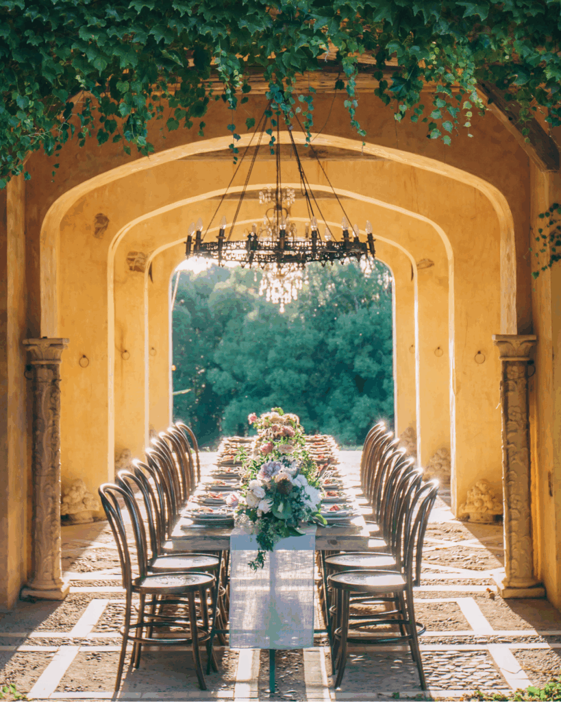 100 Breathtaking and Exclusive Australian Wedding Venues on every Couples Wish List! 17 Elegant outdoor dining setup featuring a long table adorned with floral arrangements, surrounded by arched architecture and lush greenery, creating a picturesque setting for weddings at Deux ex Belettes in Byron Bay Hinterland.