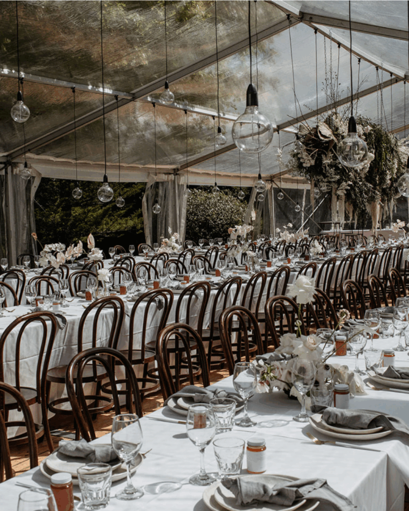 100 Breathtaking and Exclusive Australian Wedding Venues on every Couples Wish List! 19 Elegant wedding reception setup in a clear marquee with long tables adorned with white linens, glassware, and floral arrangements, featuring hanging light fixtures and a serene outdoor backdrop, ideal for intimate celebrations at a luxury venue in the Byron Bay hinterland.