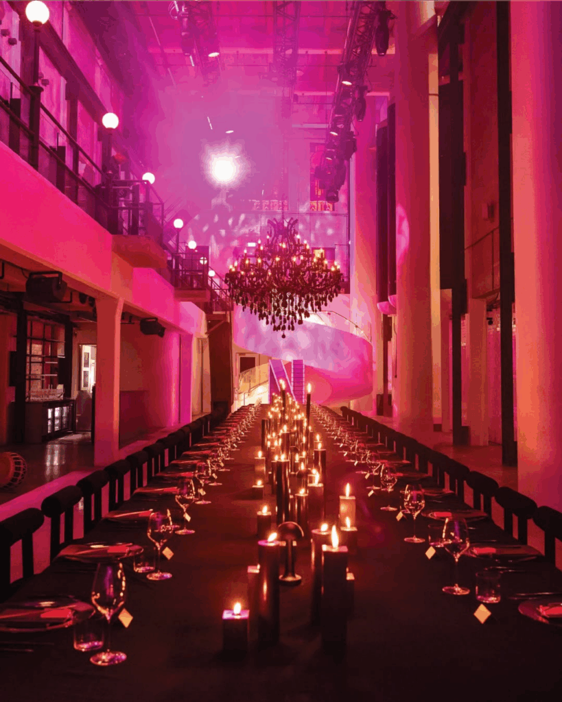 100 Breathtaking and Exclusive Australian Wedding Venues on every Couples Wish List! 37 Elegant dining setup in Machine Hall, featuring a long table adorned with candles, contemporary decor, and a grand chandelier, illuminated by soft pink lighting, ideal for wedding receptions and events.