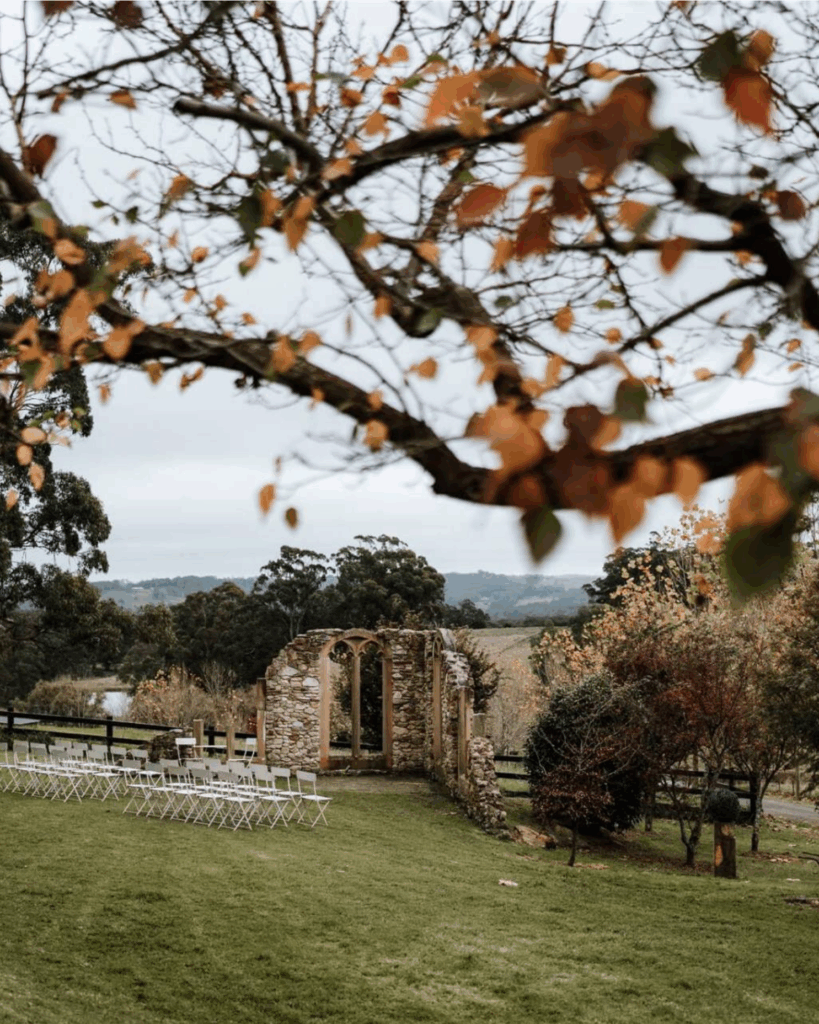 100 Breathtaking and Exclusive Australian Wedding Venues on every Couples Wish List! 56 Outdoor wedding ceremony setup at Mali Brae Farm featuring a rustic stone ruin backdrop, white folding chairs arranged on lush green grass, framed by autumn foliage and distant hills.