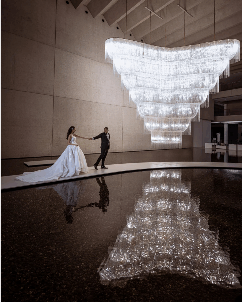 100 Breathtaking and Exclusive Australian Wedding Venues on every Couples Wish List! 63 Bride and groom holding hands in front of a stunning chandelier, reflecting in a serene water feature, showcasing a sophisticated wedding venue ambiance.