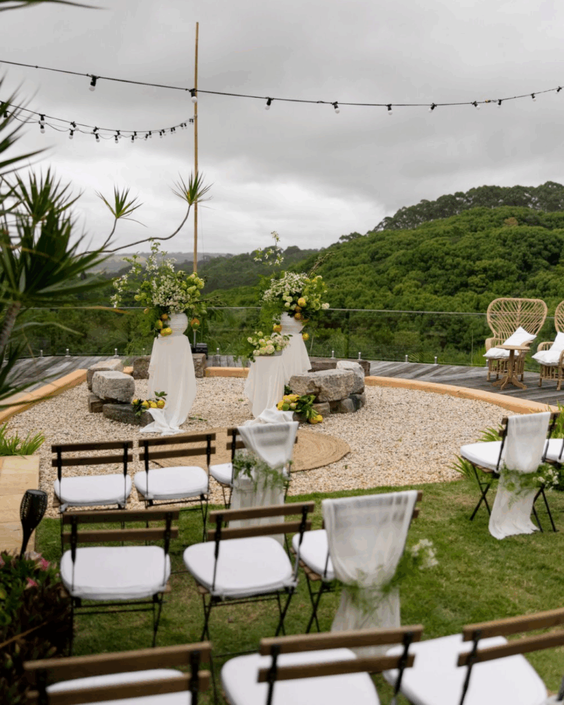 100 Breathtaking and Exclusive Australian Wedding Venues on every Couples Wish List! 20 Outdoor wedding ceremony setup at Nutting Hill Villa, featuring floral arrangements, white draped decor, and a scenic backdrop of lush greenery, with wooden chairs arranged for guests.