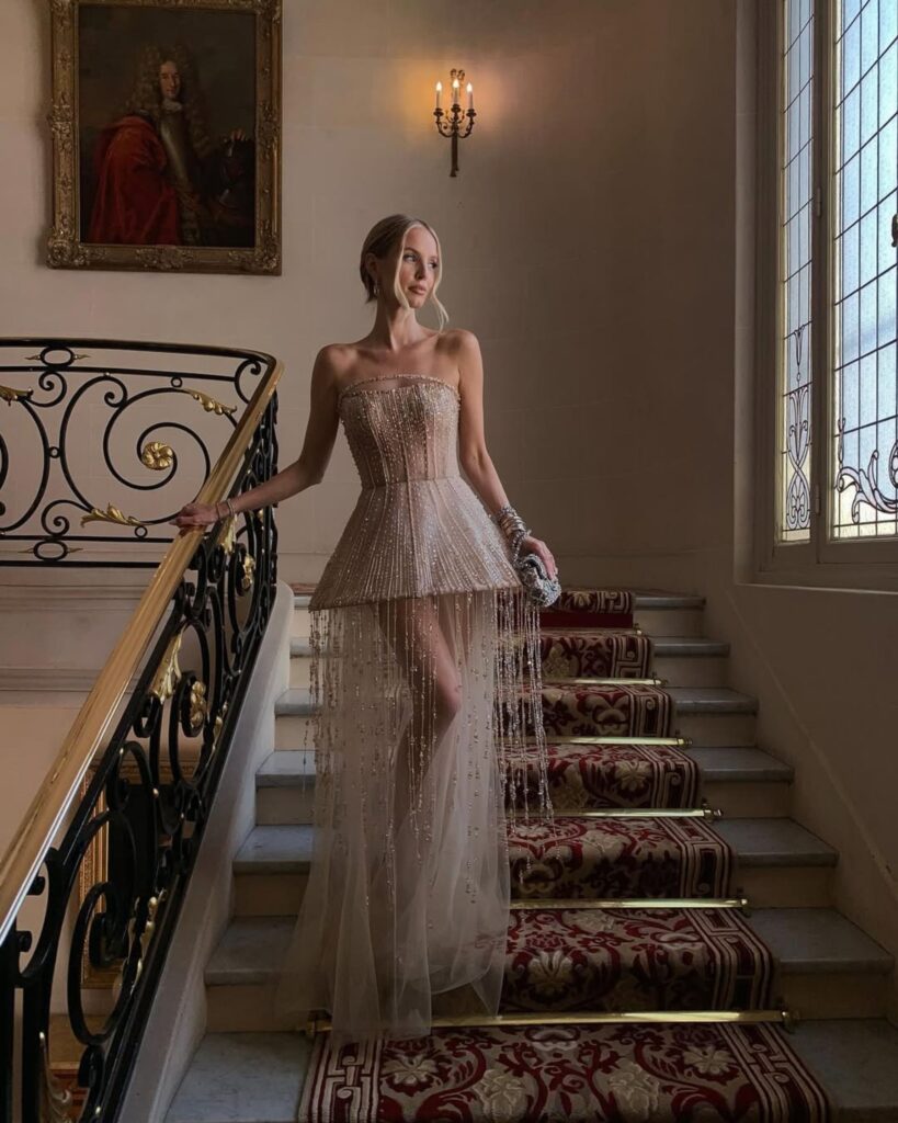 Elegant model in a beaded, couture bridal gown with a structured silhouette and flowing tulle train, posing on an ornate staircase, showcasing contemporary bridal fashion.