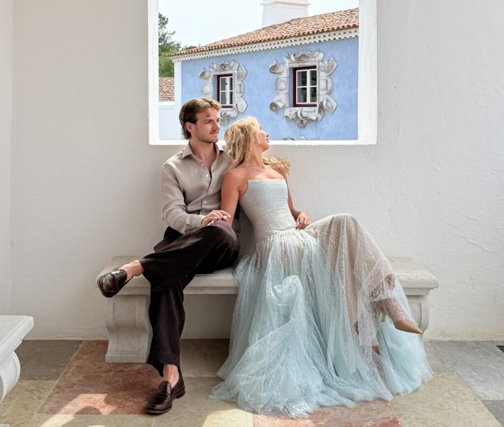 Couple sitting on a stone bench, embracing in a romantic pose, wearing elegant wedding attire, with a soft blue and white backdrop featuring architectural details, capturing an intimate moment that reflects Candid and Co.'s focus on real-time wedding storytelling.