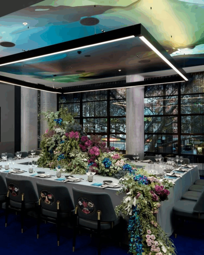 100 Breathtaking and Exclusive Australian Wedding Venues on every Couples Wish List! 40 Elegant dining setup in Studio Q at QT Sydney, featuring a long table adorned with vibrant floral arrangements, modern decor, and large windows providing natural light, ideal for intimate weddings and events.
