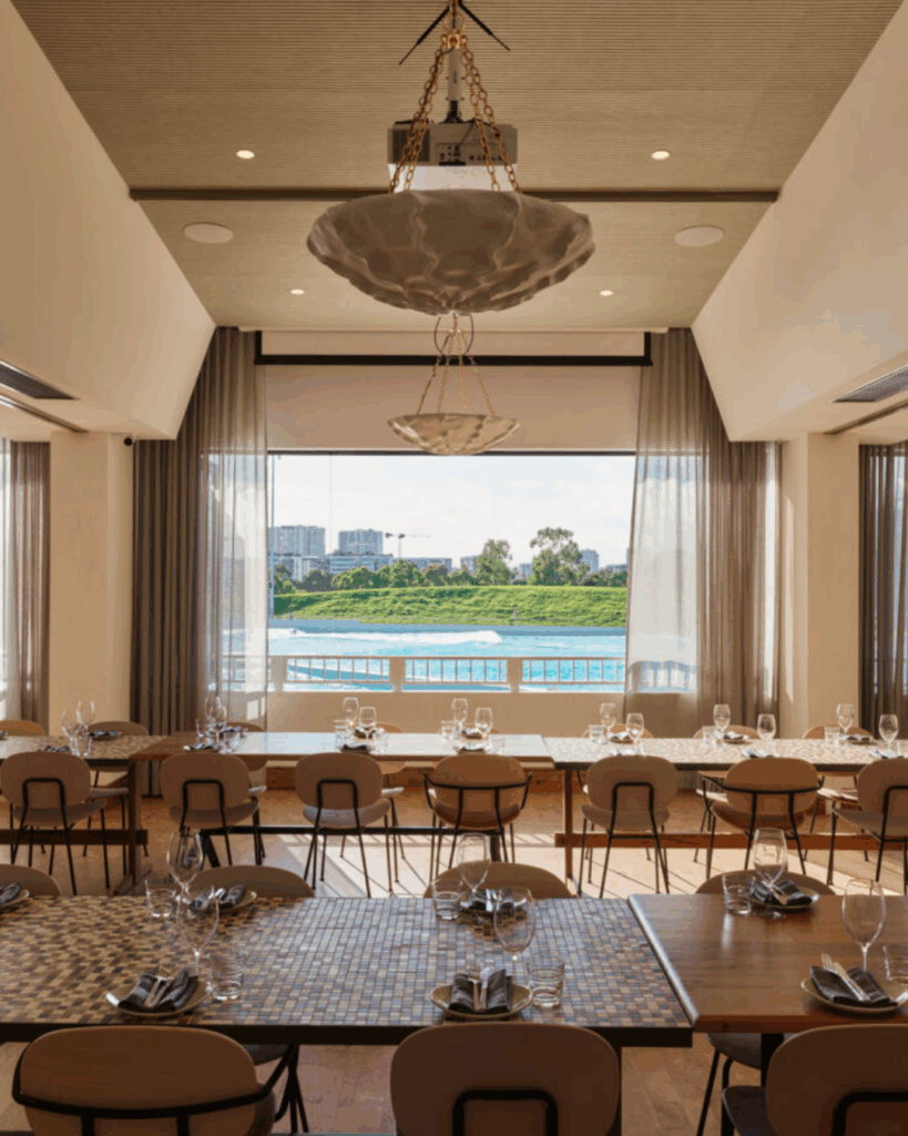 100 Breathtaking and Exclusive Australian Wedding Venues on every Couples Wish List! 41 Elegant event space at RAFI URBNSURF featuring tables set for dining, large windows showcasing views of rolling waves, modern decor with pendant lighting, suitable for private events near Sydney Olympic Park.