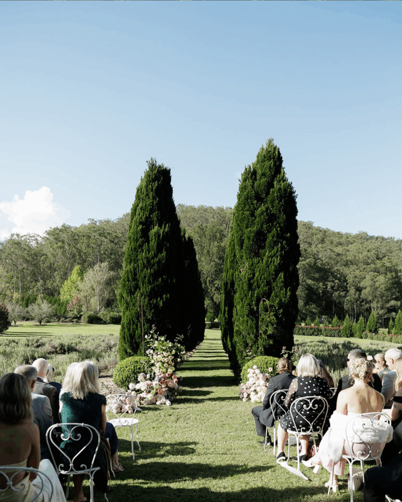 100 Breathtaking and Exclusive Australian Wedding Venues on every Couples Wish List! 12 Wedding ceremony setup in lush garden at The Redleaf Wollombi, featuring guests seated on elegant chairs, flanked by tall cypress trees and floral arrangements along the aisle.