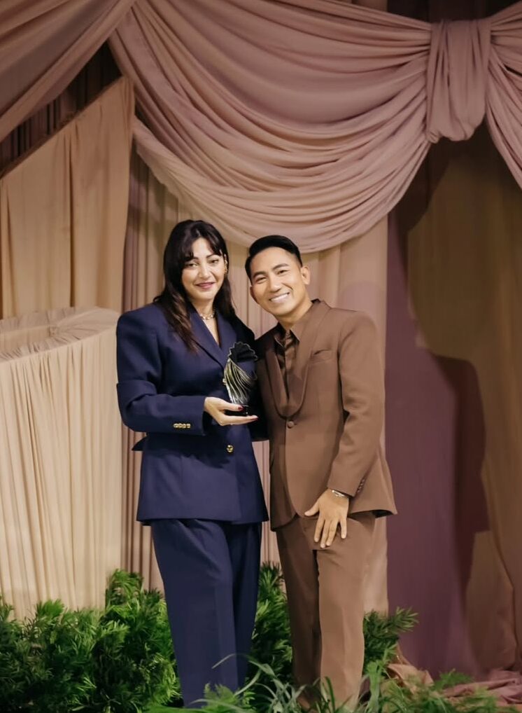 Woman in navy suit and man in brown suit holding an award on stage, celebrating achievements in the wedding industry at Gideon Hermosa’s Gala Night Extravaganza.