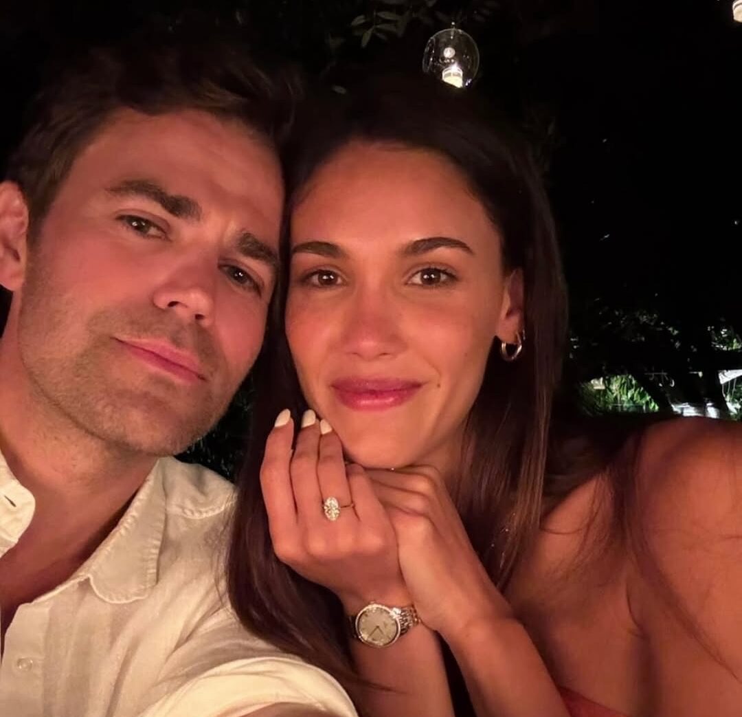 Paul Wesley and Natalie Kuckenburg smiling together, showcasing her engagement ring and a romantic setting, highlighting their engagement news.