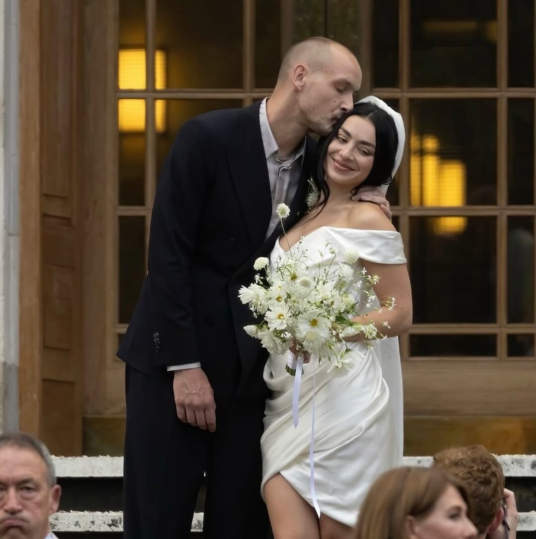 Bride in off-the-shoulder white wedding dress holding bouquet of white flowers, smiling with groom in black suit kissing her forehead, celebrating their wedding outdoors.