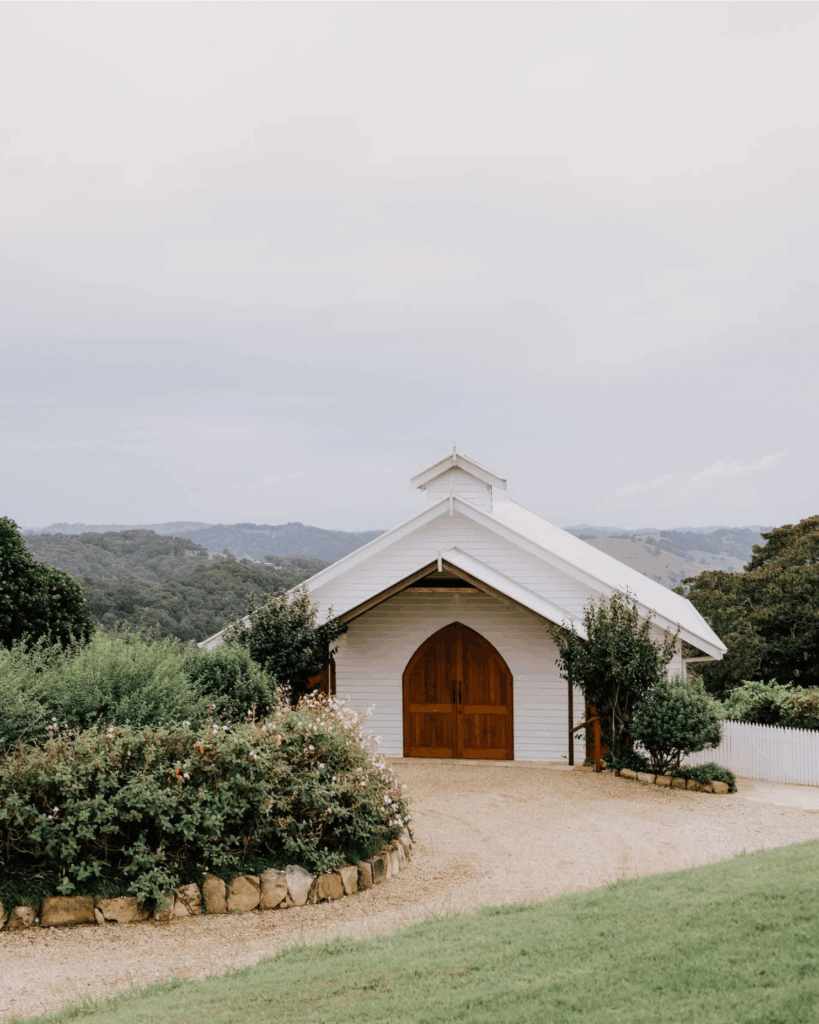 100 Breathtaking and Exclusive Australian Wedding Venues on every Couples Wish List! 21 White chapel surrounded by lush greenery and a gravel pathway, set against a scenic backdrop of hills in the Byron Bay hinterland, ideal for wedding ceremonies at exclusive venues like Nutting Hill Villa and Victoria’s Estate.