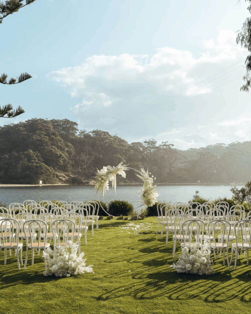 100 Breathtaking and Exclusive Australian Wedding Venues on every Couples Wish List! 15 Outdoor wedding setup at The Cove in Jervis Bay, featuring white chairs arranged on lush green grass, a floral arch, and a serene water backdrop, ideal for romantic ceremonies.