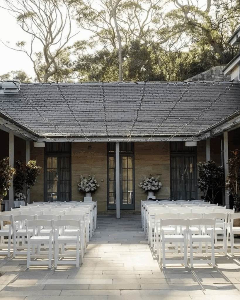 100 Breathtaking and Exclusive Australian Wedding Venues on every Couples Wish List! 45 Outdoor wedding ceremony setup at the Gunners Barracks featuring white chairs arranged in rows, surrounded by greenery and elegant floral arrangements, showcasing a blend of heritage and contemporary design.