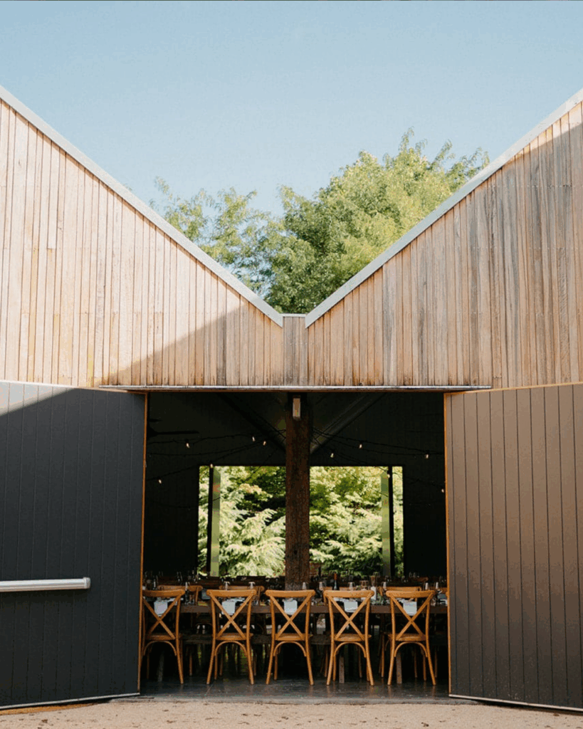 100 Breathtaking and Exclusive Australian Wedding Venues on every Couples Wish List! 26 Barn-style reception space at The Woodhouse Wollombi, featuring wooden architecture, filled with rustic dining tables and surrounded by lush greenery, ideal for weddings in the Hunter Valley.