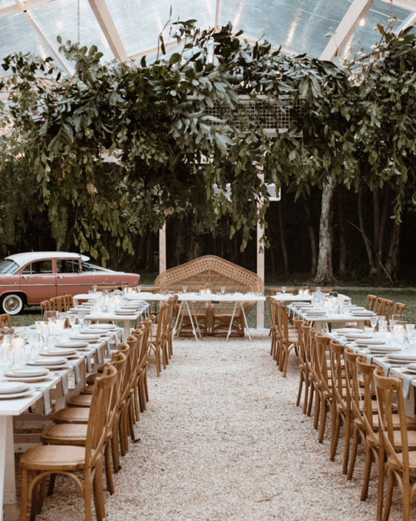 100 Breathtaking and Exclusive Australian Wedding Venues on every Couples Wish List! 22 Elegant outdoor dining setup under a clear roof pavilion at Victoria’s Estate in Byron Bay, featuring long tables adorned with plates and glassware, surrounded by lush greenery and a vintage car in the background.