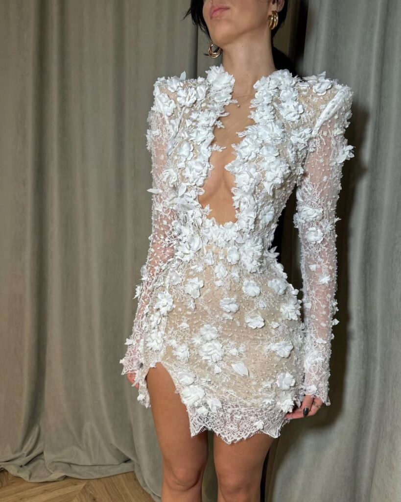 Model showcasing a luxurious, handcrafted white gown adorned with intricate floral embellishments and a daring neckline, set against a soft, neutral backdrop.