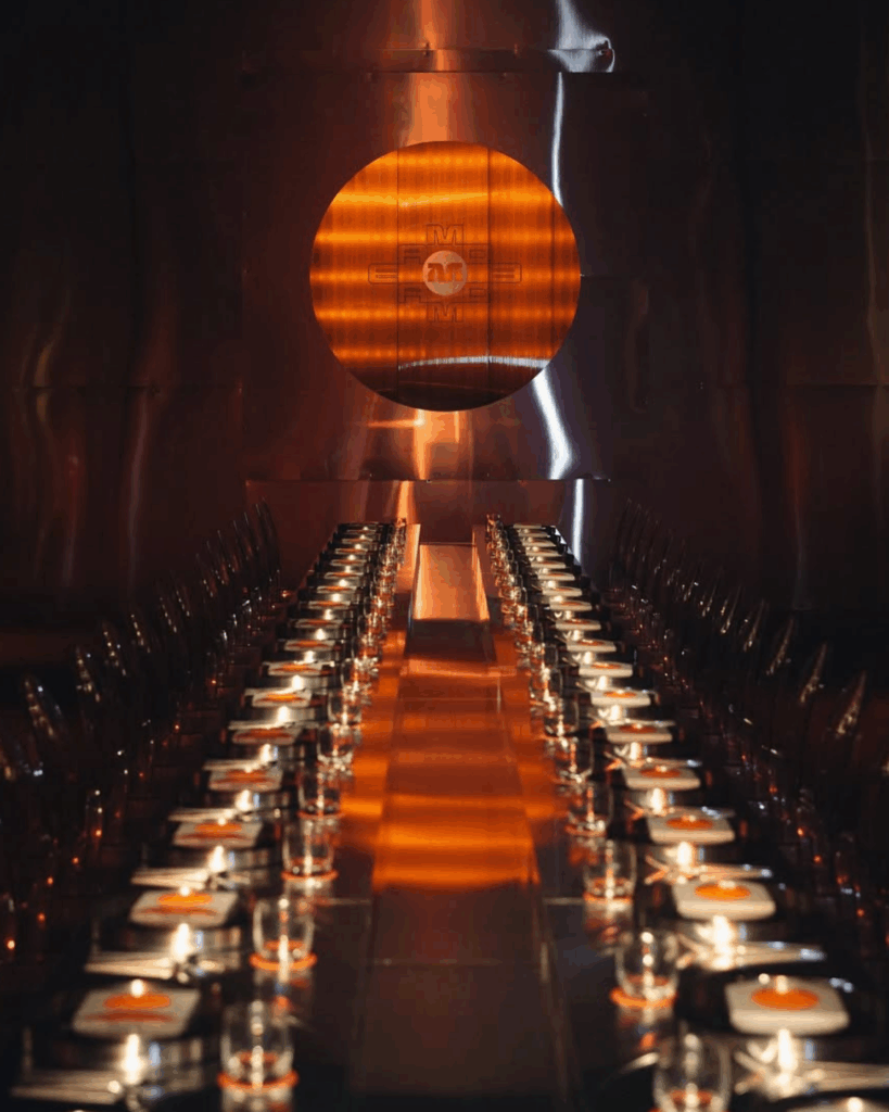 100 Breathtaking and Exclusive Australian Wedding Venues on every Couples Wish List! 39 Elegant long dining table set for an event, featuring illuminated candles and contemporary design elements, against a backdrop of polished metal with an orange circular feature, reflecting a stylish venue atmosphere suitable for weddings and celebrations.