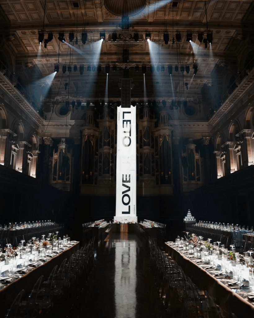 100 Breathtaking and Exclusive Australian Wedding Venues on every Couples Wish List! 43 Elegant wedding reception setup in a grand hall featuring long tables with glassware and floral arrangements, illuminated by dramatic lighting, showcasing a central banner with "LOVE" and "LETO" in a historic venue.